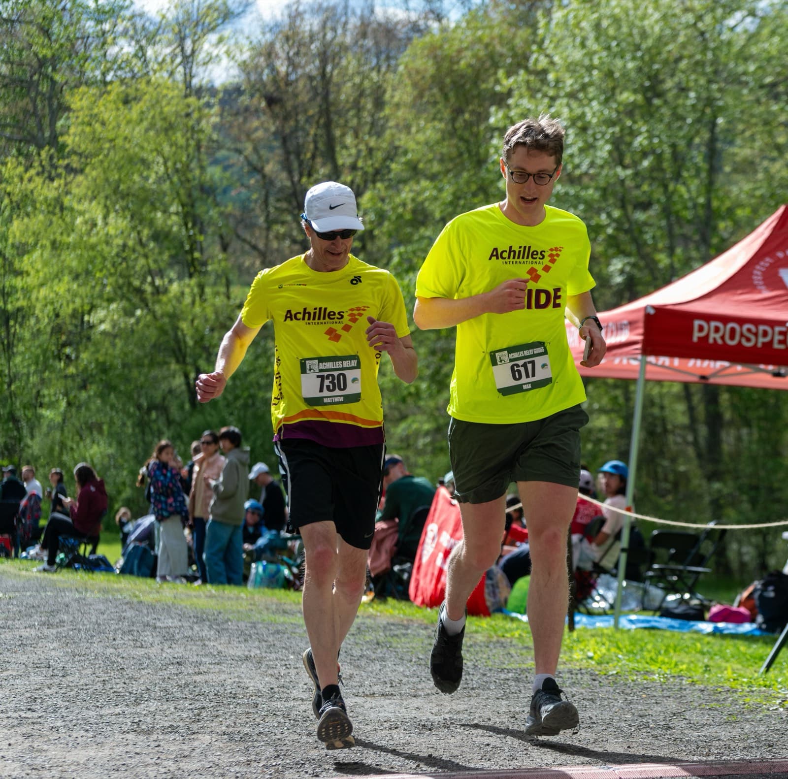 Max, in an Achilles International guide shirt, running a race on a gravel path alongside another runner in an Achilles shirt.